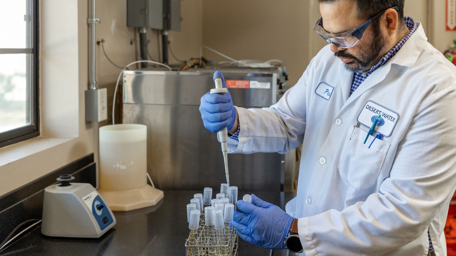 A scientist in a lab coat uses a pipette to transfer liquid into test tubes, surrounded by laboratory equipment.