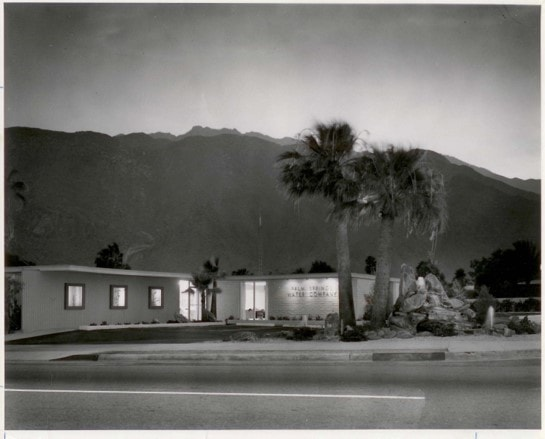 A black-and-white photo of a building with palm trees in front, set against a mountainous backdrop.