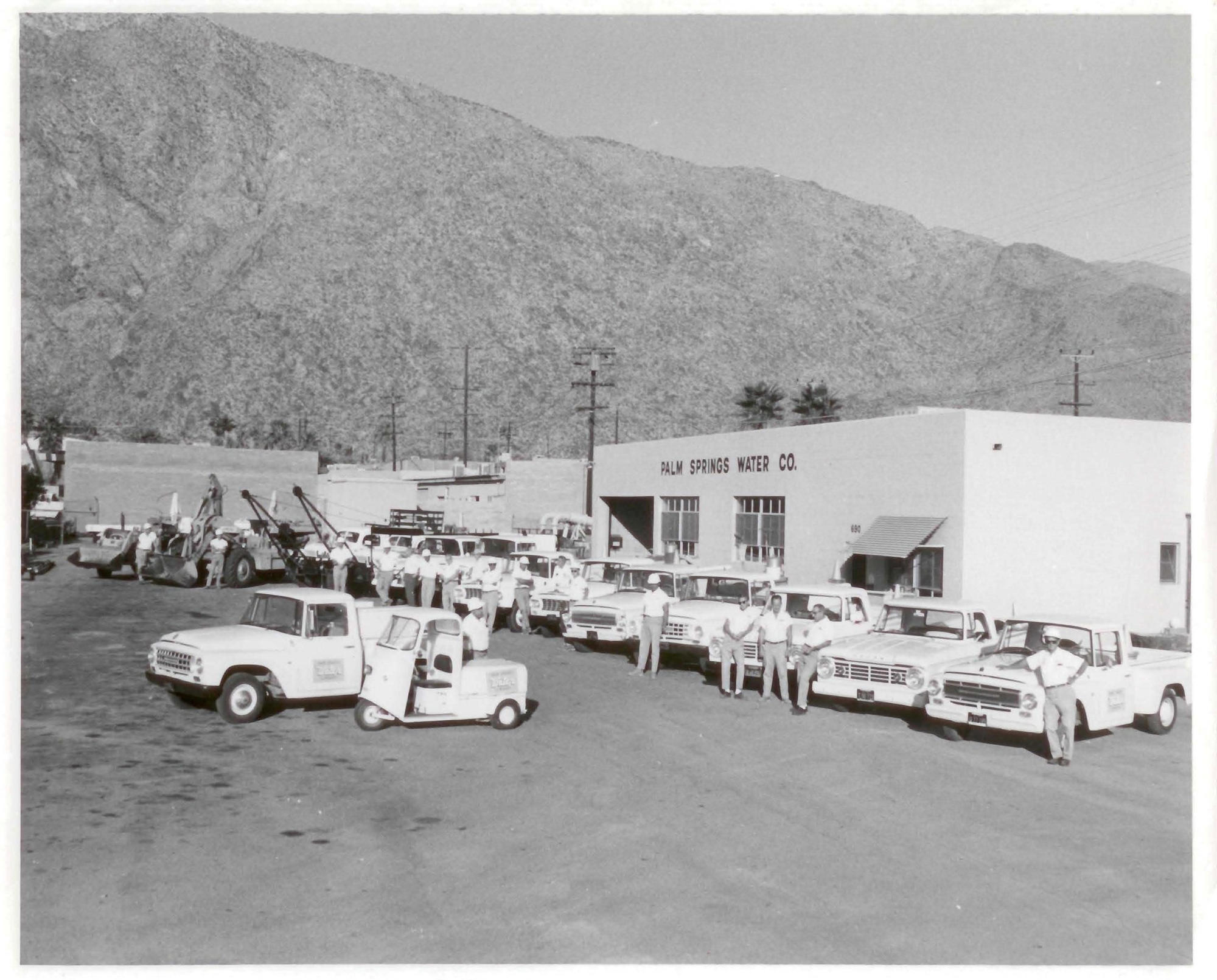 A black-and-white photo of Palm Springs Water Co. employees lined up with their vehicles in front of a building and mountains.