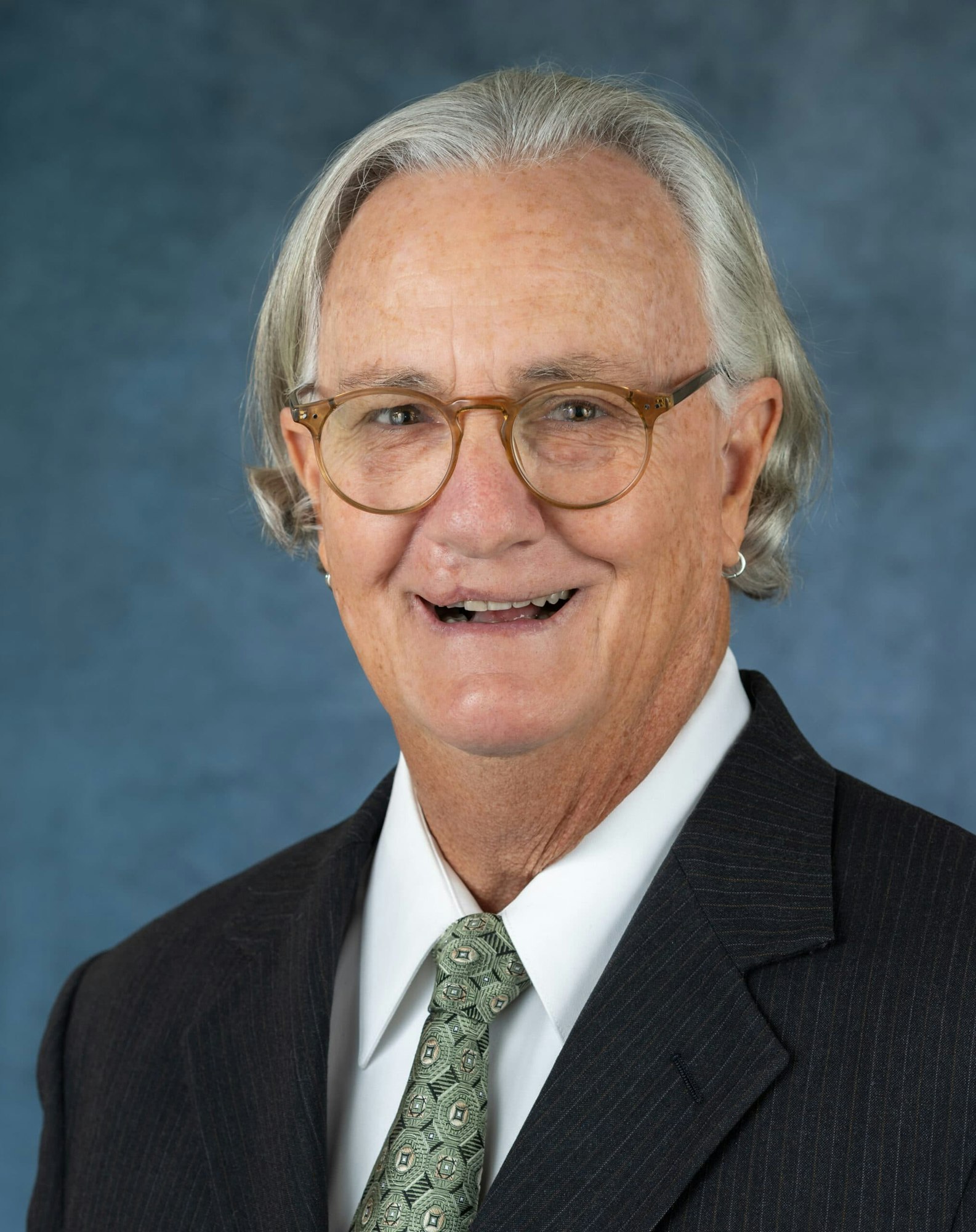 A smiling man with long gray hair, wearing glasses and a suit with a patterned tie, against a blue background.