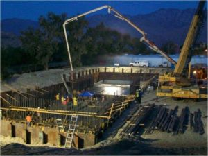 A construction site at night with workers, a concrete pump, and a foundation being poured into a form structure.