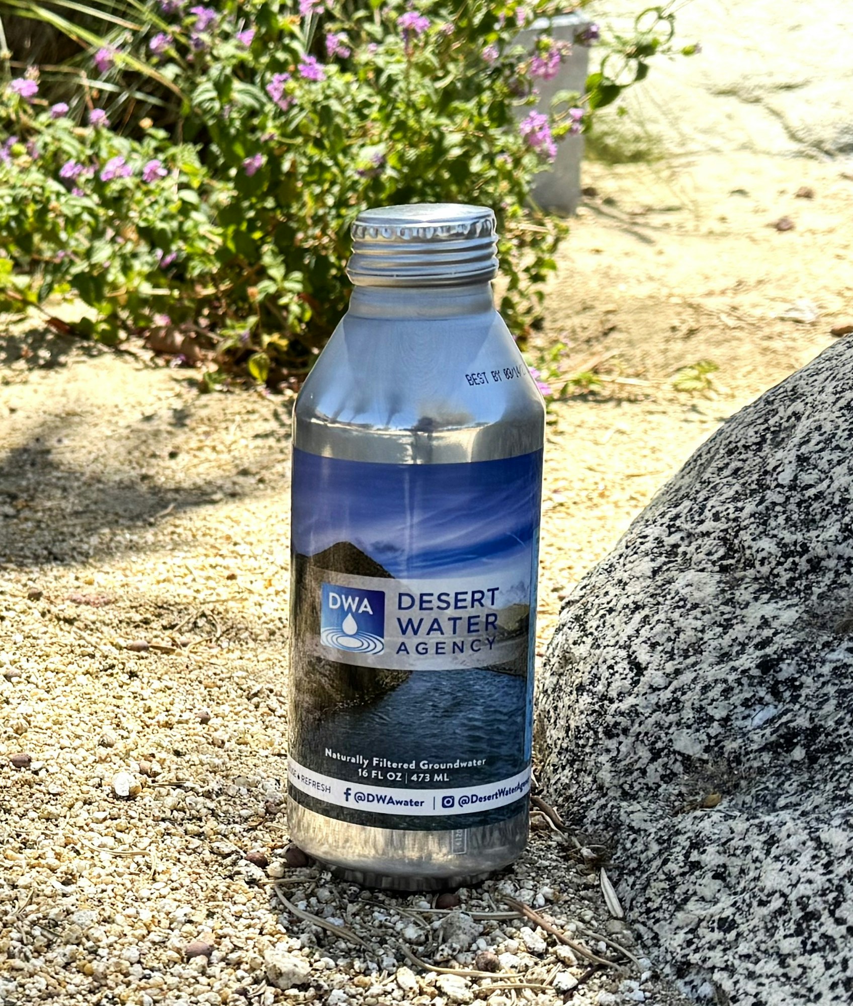 A metallic water bottle from the Desert Water Agency featuring naturally filtered groundwater, set against a sandy background.