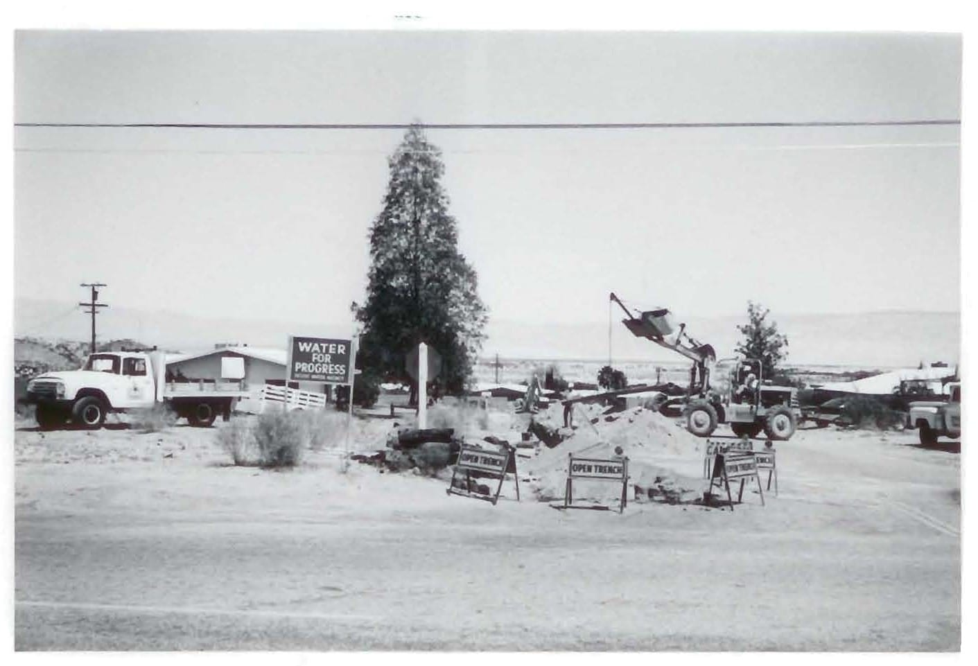 A black-and-white image of a construction site with trucks, a sign that says "Water for Progress," and excavation activity.