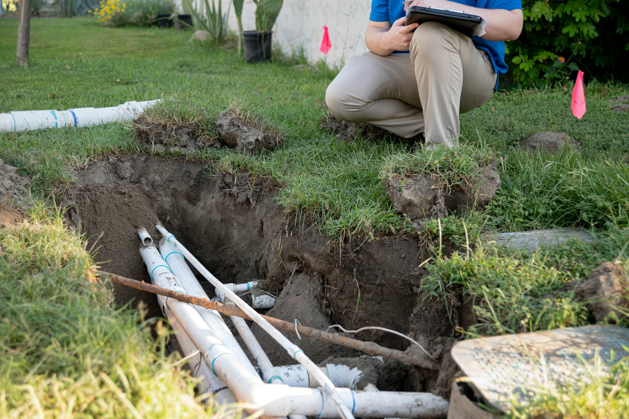 A person crouches by a trench in grass, inspecting exposed PVC pipes, possibly for plumbing or irrigation work.