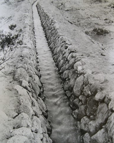 The image features a narrow, water-filled irrigation canal lined with stones, surrounded by bare earth and sparse vegetation.