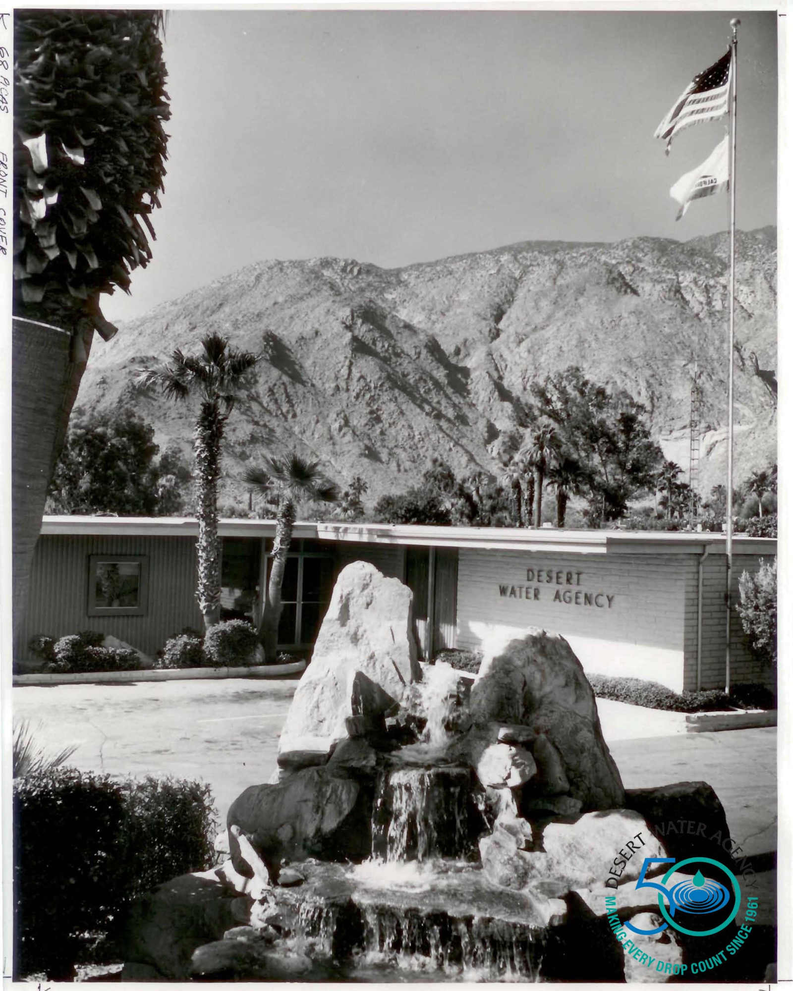 A black-and-white photo of the Desert Water Agency building, featuring a rock fountain and palm trees, with mountains in the background.