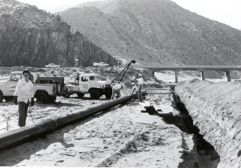 The image shows workers installing a large pipe in a sandy area, with vehicles and a bridge in the background, captured in black and white.