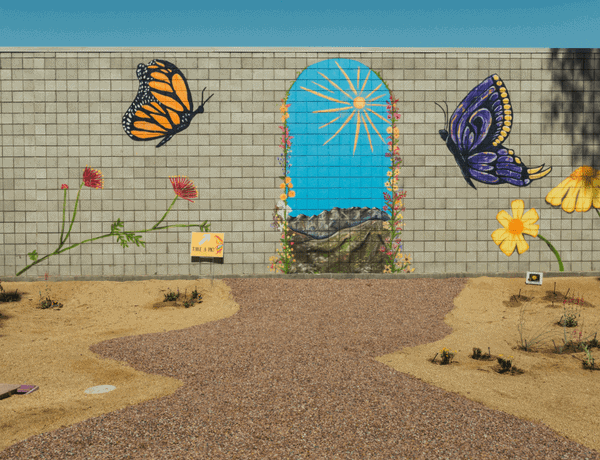 A vibrant mural featuring butterflies and an archway with a scenic backdrop, set in a landscaped area with a pathway.