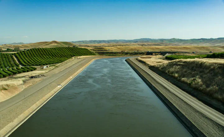 A serene landscape featuring a wide water canal bordered by rows of lush greenery and rolling hills under a clear blue sky.