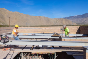 Two workers in safety gear are installing steel beams on a building structure, with mountains in the background.