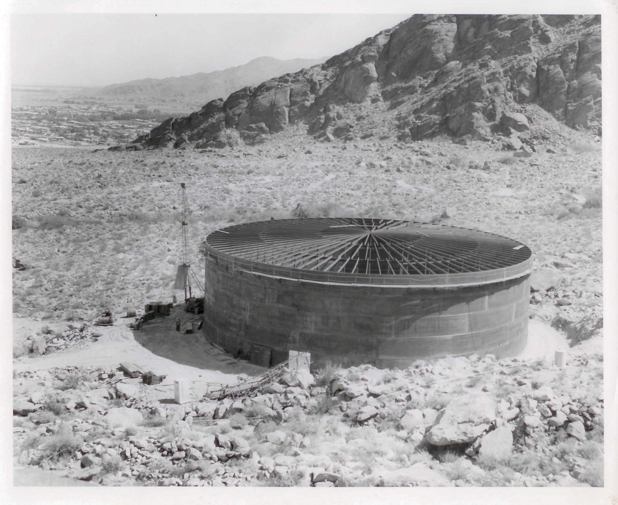 A large circular concrete structure sits in a rocky desert landscape, surrounded by mountains and sparse vegetation.
