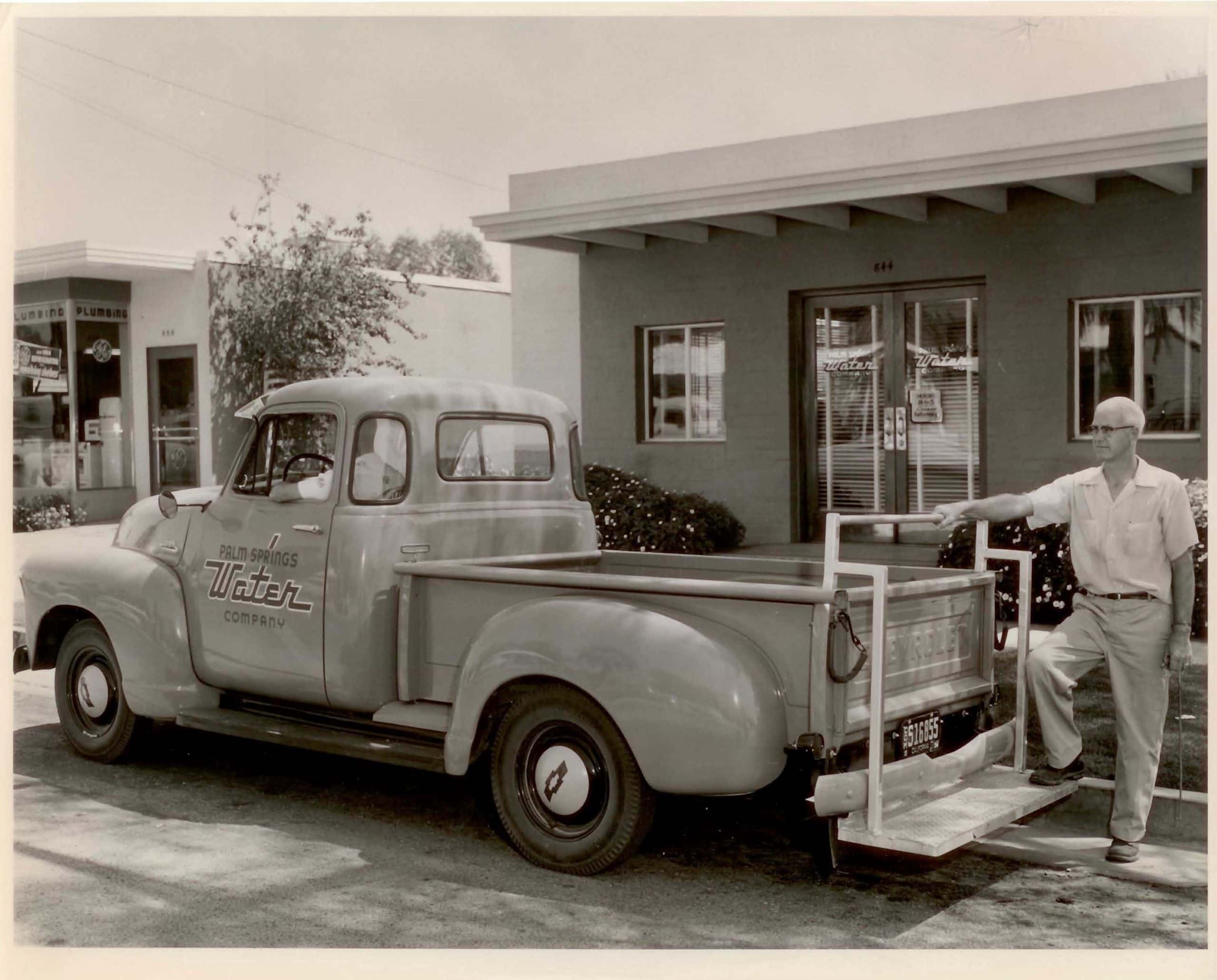 A vintage photo of a man beside a classic truck labeled "Palm Springs Water Company," in front of a building.