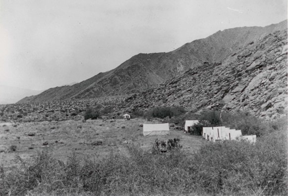 A black-and-white photo of a mountainous landscape with several tents set up in a sparse, open area.