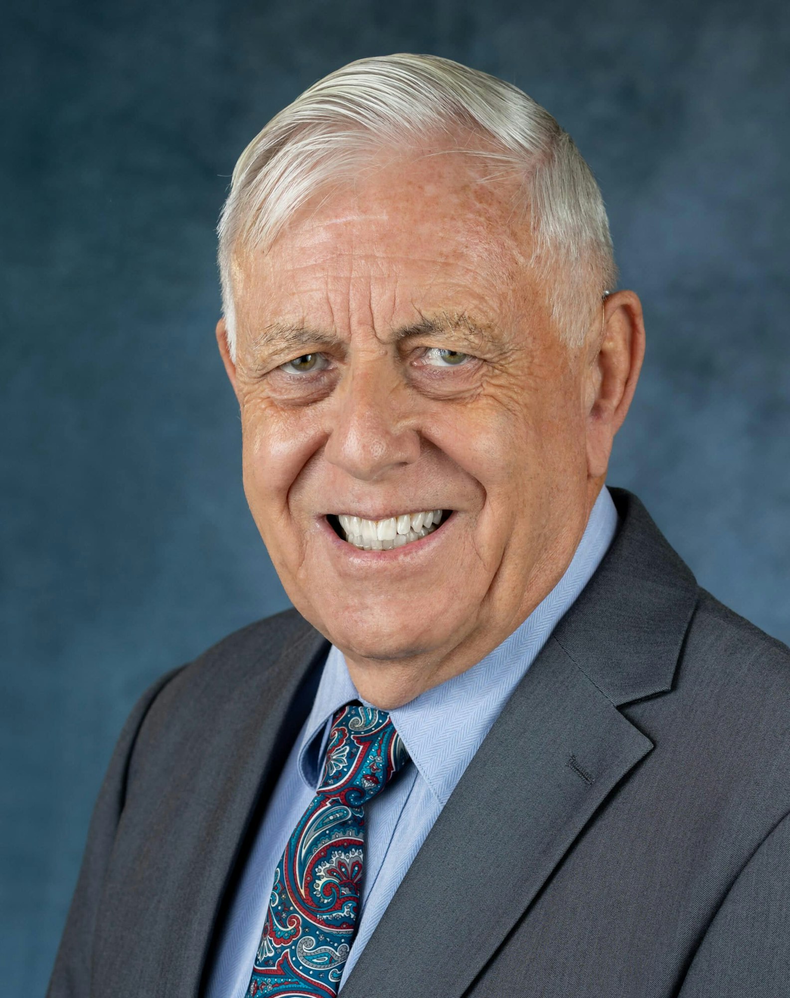 The image shows an older man smiling, wearing a dark suit and a colorful patterned tie, with a blue backdrop.