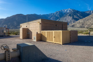 A small building with wooden fencing, set against mountainous terrain and clear blue skies. Gravel surrounds the structure.