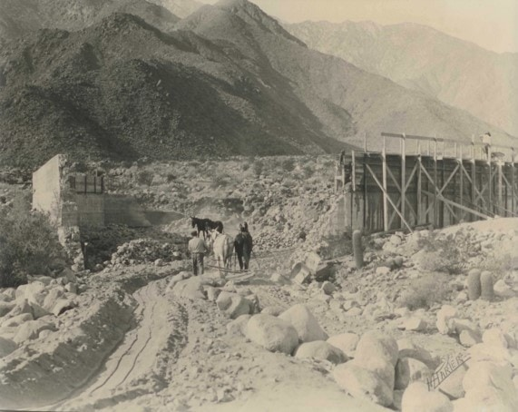 A historical scene featuring people walking along a rocky path, mountains in the background, and a wooden structure nearby.