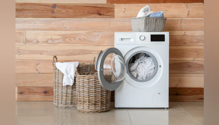 A modern white washing machine with an open door, surrounded by woven baskets filled with laundry, against a wooden wall.