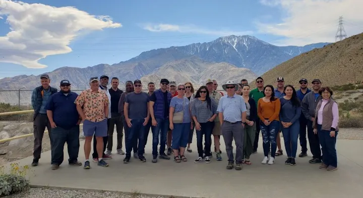 A group of people posing for a photo with mountains in the background on a sunny day.