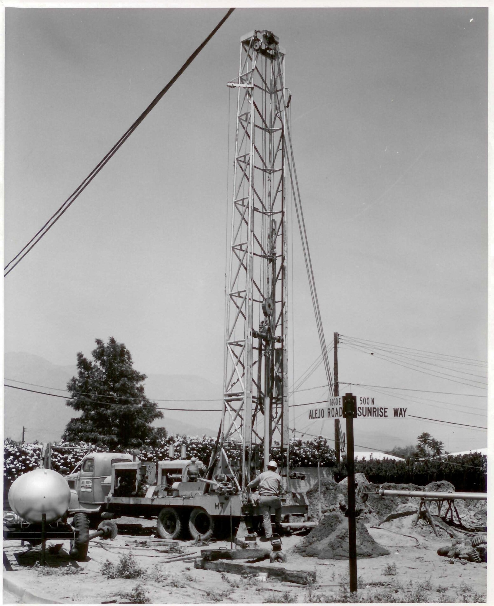 A black-and-white image of a drilling rig with workers, a truck, and street signs, set in an outdoor area with trees.