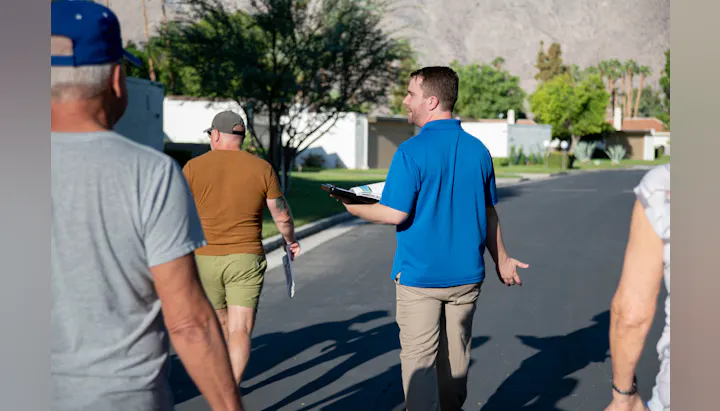 A group of people is walking on a sunny street, with one person in a blue shirt holding a clipboard and talking.