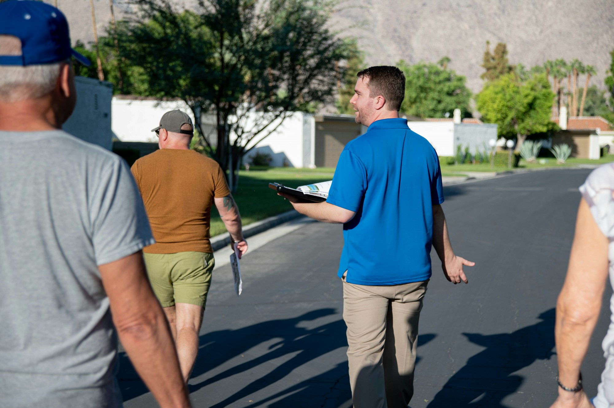 A group of people is walking on a sunny street, with one person in a blue shirt holding a clipboard and talking.