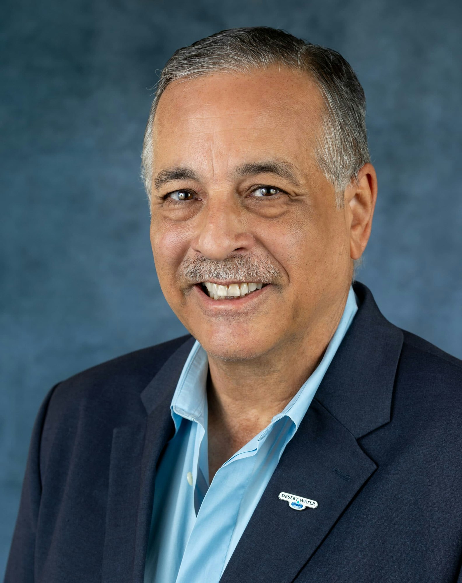 A smiling man wearing a blue blazer and light blue shirt, with a pin labeled "DESERT WATER" against a blue backdrop.