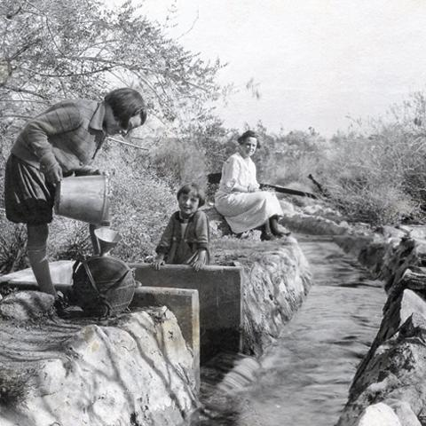 The image shows two women and a child by a stream, one pouring water while another sits nearby, all in a natural setting.