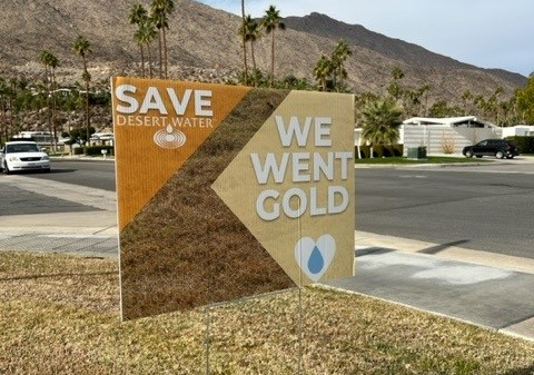 A yard sign promoting water conservation with the message "SAVE DESERT WATER" and "WE WENT GOLD" displayed against a desert backdrop.