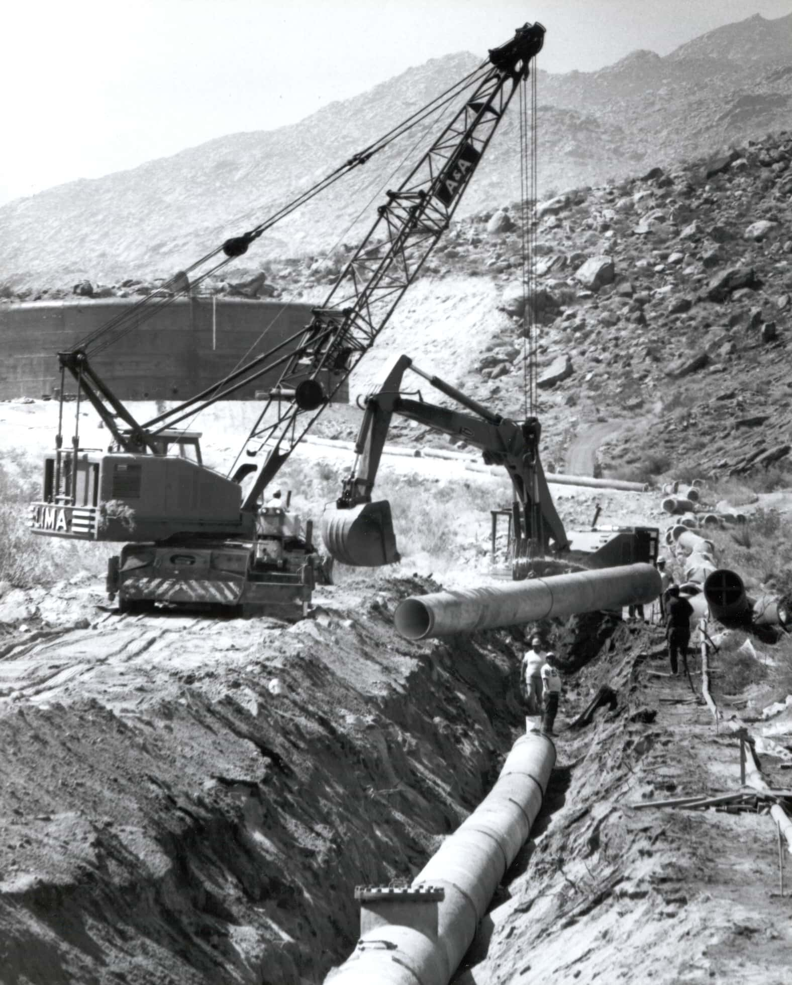 A construction site with cranes and workers laying large pipes in a trench, surrounded by mountainous terrain.