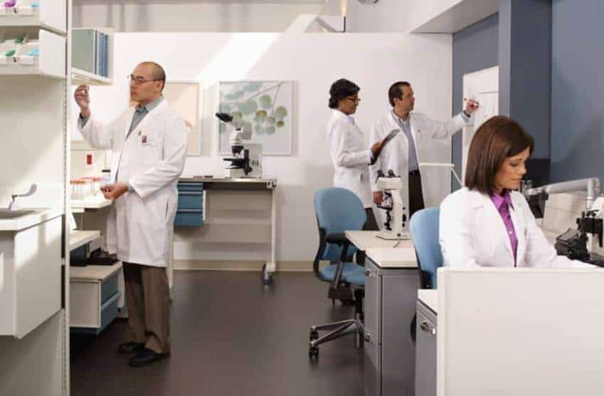 A laboratory setting featuring four individuals in lab coats engaged in various tasks, including using equipment and discussing findings.