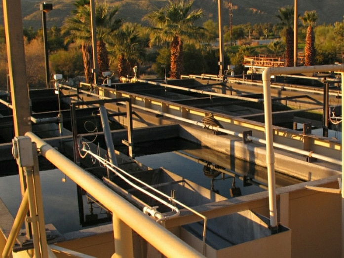 The image shows a water treatment facility with tanks, pipes, and palm trees in the background, under natural lighting.