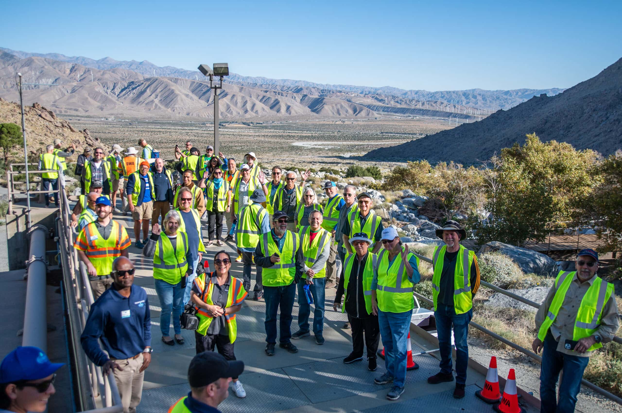 A large group of people wearing bright yellow safety vests pose for a photo against a scenic mountainous backdrop.