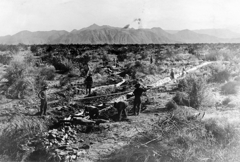 Workers are seen constructing infrastructure in a barren landscape with mountains in the background, possibly involving irrigation or railroads.