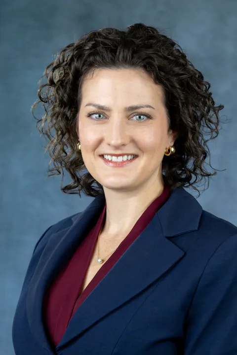 A smiling woman with curly hair wearing a navy blazer and a maroon top, positioned against a textured blue background.