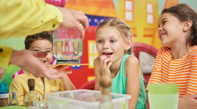 Children are excitedly watching a science experiment involving a glass of water, led by an adult.