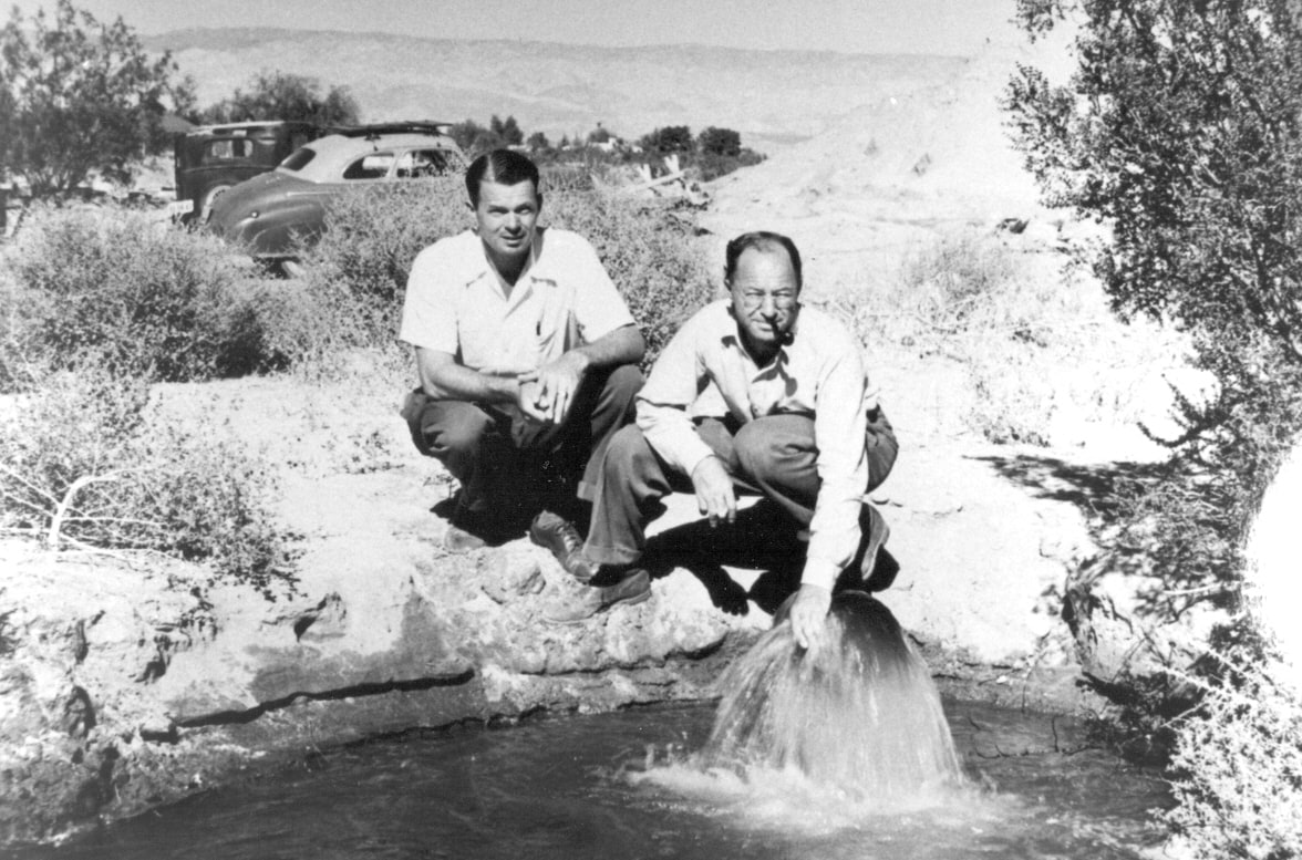 Two men kneel by a water spring in a desolate area, with mountains in the background and vintage cars visible. Black and white photo.