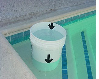 A bucket partially submerged in water, with arrows indicating water level, likely used for measuring or testing pool water.