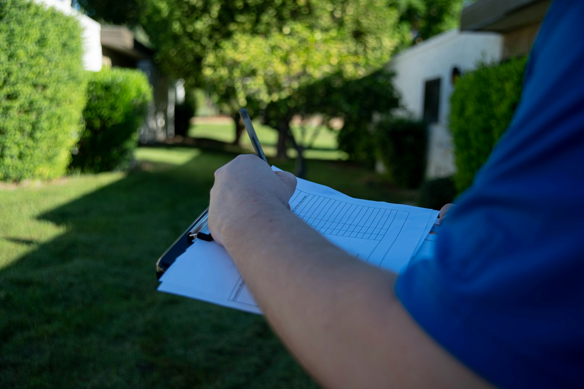 A person is holding a clipboard with a pen, writing notes outside in a green yard with trees and plants around.