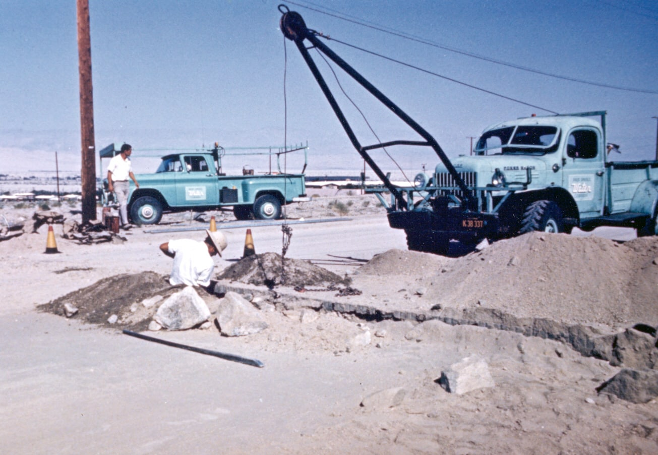 A construction site with two vintage trucks and a worker digging, surrounded by piles of dirt and rocks.