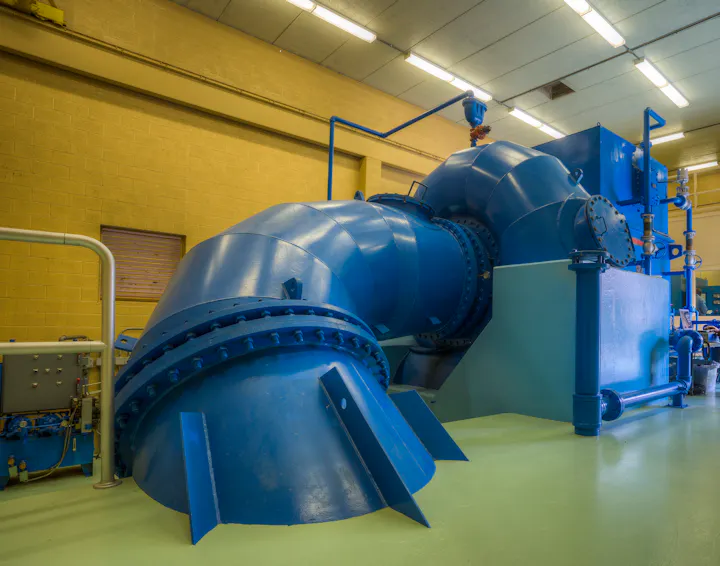 The image shows a large blue industrial pump or turbine system within a facility, featuring pipes and control equipment.