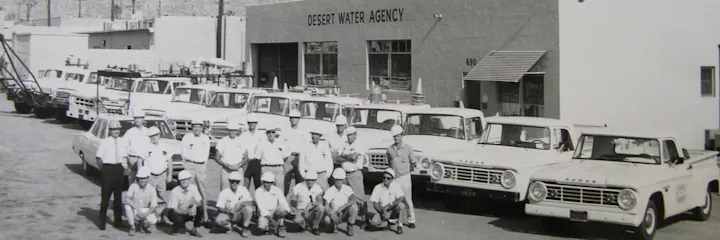 A historic black-and-white photo of the Desert Water Agency, featuring staff members and several trucks lined up outside.