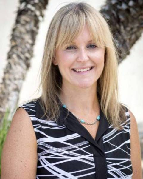 A smiling woman with blonde hair in a patterned shirt stands outdoors, with palm trees in the background.