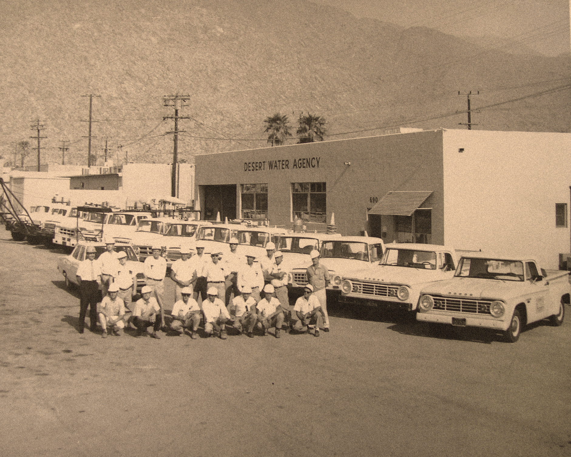 A historic photo of the Desert Water Agency staff with several trucks parked outside their building, set against a mountainous backdrop.