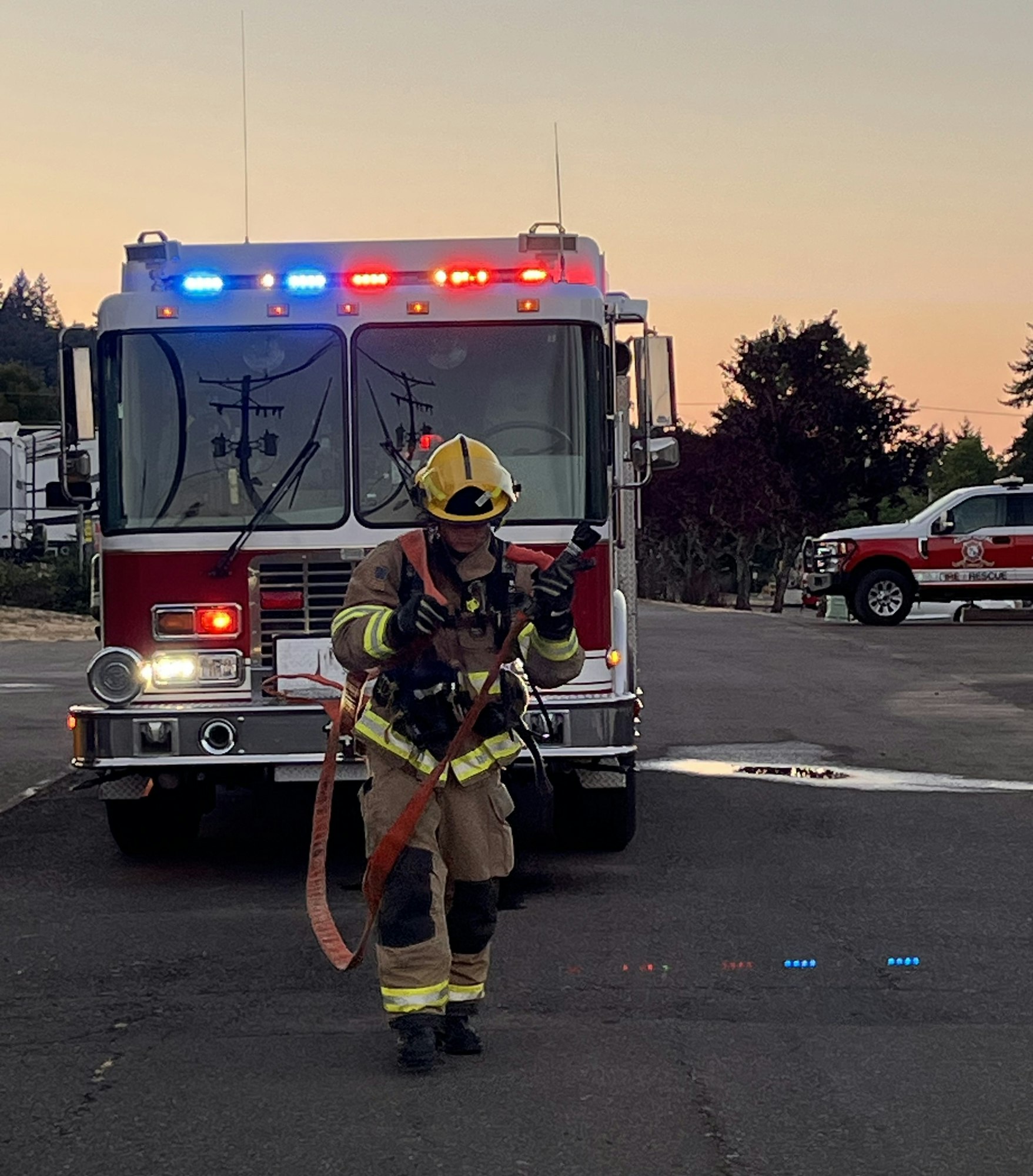A firefighter in gear walks toward a fire truck with emergency lights flashing at sunset, ready for action.