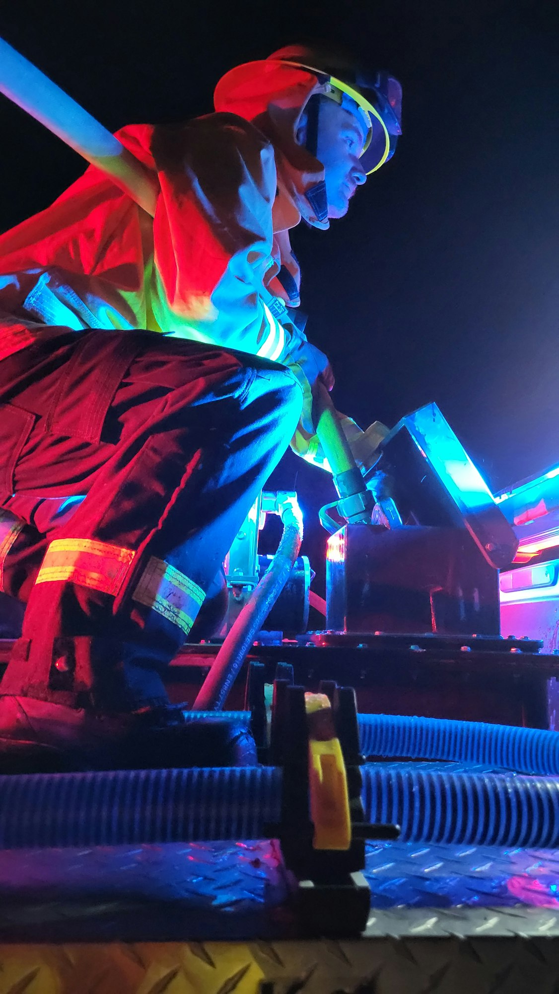 A person in firefighting gear is working at night, illuminated by colorful lights, possibly on a fire truck or equipment.