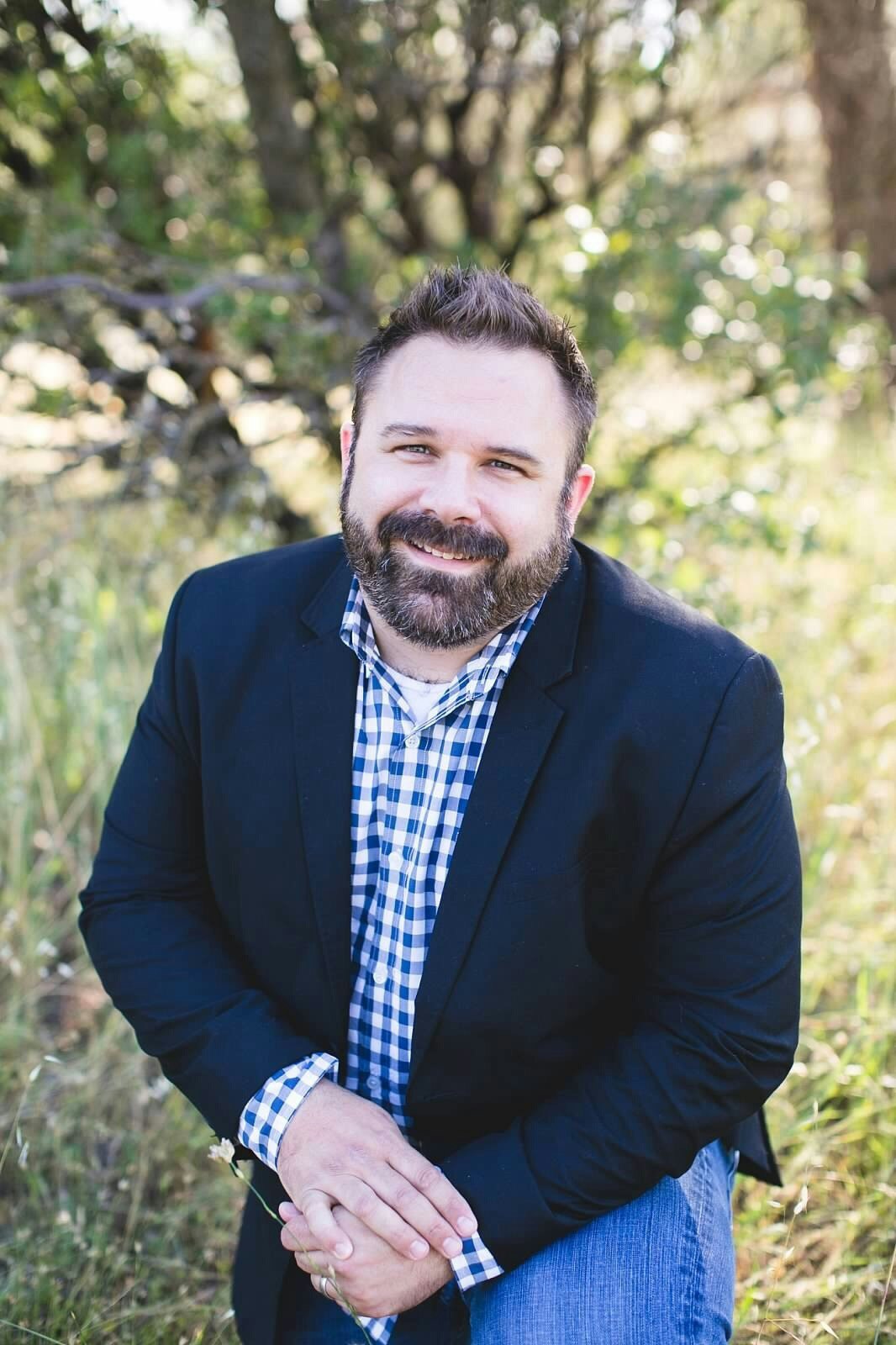 A smiling man in a blazer over a gingham shirt, hands clasped, standing in a sunny, wooded area.