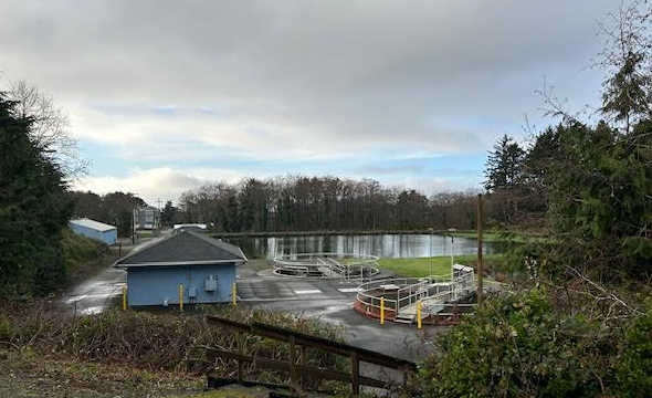 A water treatment facility with circular tanks, a small building, and surrounding greenery under a cloudy sky.