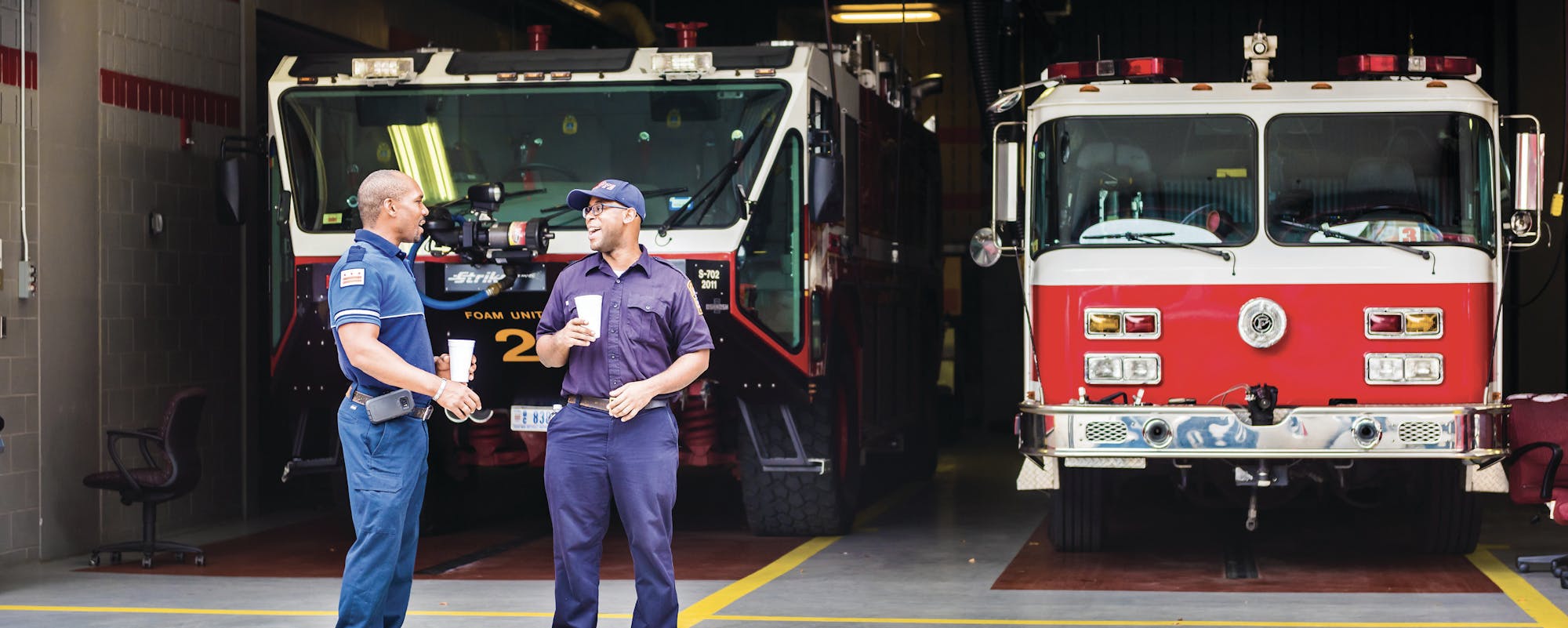 Two firefighters in conversation, holding cups in front of two fire trucks in a fire station garage.