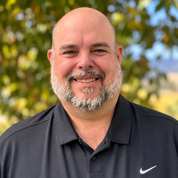 A smiling man with a beard wearing a black polo shirt stands outdoors with greenery in the background.