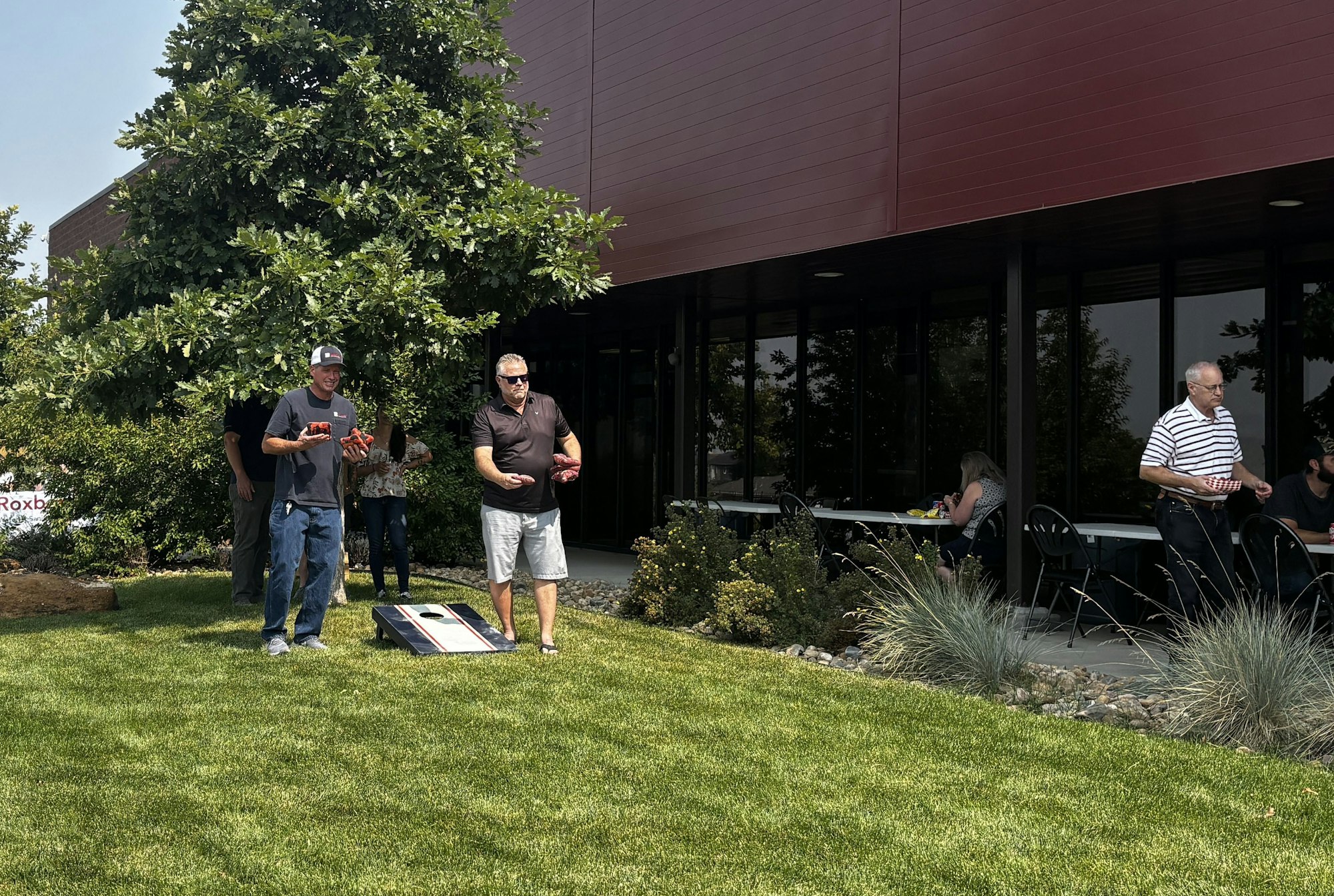People playing cornhole outside near a building, with more people seated at tables.